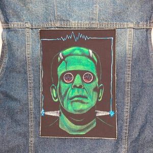May include: A black and green Frankenstein monster patch with a blue background. The monster is wearing glasses with a blue and white design. The patch has a black border and is sewn onto a denim jacket.
