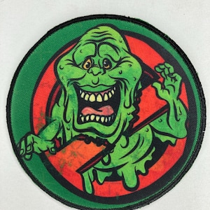 Puede incluir: Un parche circular verde y rojo con una imagen de dibujos animados de un fantasma verde, viscoso y sonriente con una boca grande y dientes afilados. El fantasma gotea baba verde y tiene un fondo rojo.