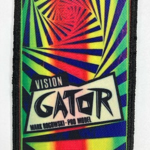 Puede incluir: Parche rectangular con un diseño psicodélico vibrante. El parche presenta un patrón arremolinado de rojo, amarillo, azul y verde, con el texto "VISION GATOR" y "MARK ROGOWSKI - PRO MODEL" en negrita. El parche está bordeado con costuras negras.