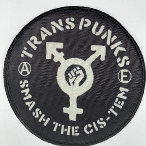 May include: A circular black embroidered patch featuring white text that reads TRANS PUNKS SMASH THE CIS-TEM. The centre displays a white transgender symbol with a raised fist icon inside the circle, flanked by an anarchy symbol and an E symbol.