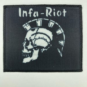 Può includere: Una toppa rettangolare in tessuto nero con una grafica serigrafata bianca di un teschio umano con un'acconciatura mohawk a punte. Il testo Infa-Riot è stampato in bianco sopra il disegno del teschio. La toppa ha un bordo nero cucito.