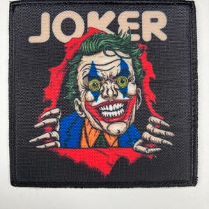 Op de afbeelding: Een vierkante geborduurde patch met een afbeelding van de Joker met groen haar, blauwe ruitvormige oogmake-up en een brede grijns. Het personage scheurt door een zwarte achtergrond, met het woord JOKER in beige bovenaan gedrukt.