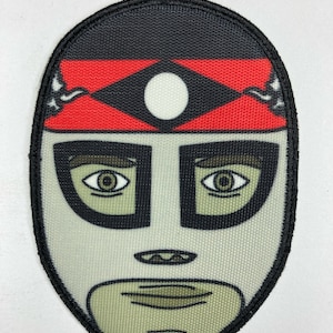 Puede incluir: Un parche bordado en blanco y negro de un luchador enmascarado con una cinta roja en la cabeza y un círculo blanco en la frente.