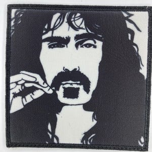 Pode incluir: Patch em preto e branco com um retrato estilizado de um homem com cabelo comprido, bigode e cavanhaque. O patch tem uma borda preta e um fundo branco. O homem está a tocar no bigode com os dedos.