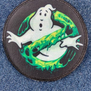 Puede incluir: Un parche bordado circular que presenta un personaje fantasma blanco con una expresión de sorpresa, rodeado por un gráfico de baba verde vibrante. El diseño está sobre un fondo negro con un borde cosido, creando un aspecto de alto contraste.