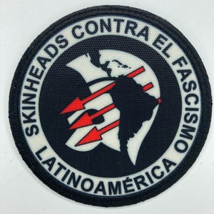 Puede incluir: Parche redondo con borde y fondo negros. El texto blanco dice "SKINHEADS CONTRA EL FASCISMO LATINOAMERICA". Un contorno blanco de las Américas está en el centro, con tres flechas rojas que apuntan a Sudamérica.