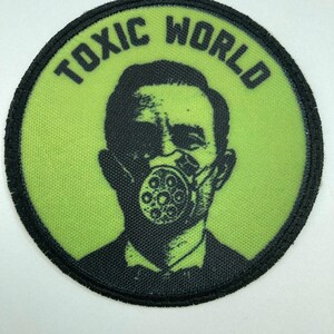 May include: A circular embroidered patch with a lime green background and a black border. The patch features a black graphic illustration of a person wearing a gas mask, with the text TOXIC WORLD printed in black capital letters along the top edge.