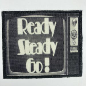 Puede incluir: Un parche bordado en blanco y negro con una pantalla de televisión vintage que muestra las palabras "Ready Steady Go!"