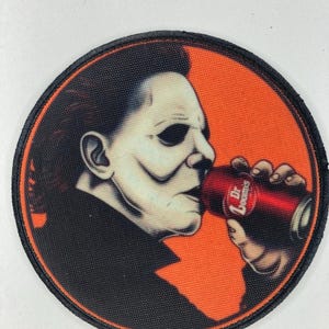 Puede incluir: Parche redondo que representa a un personaje bebiendo de una lata de refresco Dr. Loomis. El personaje está representado en negro, blanco y gris sobre un fondo naranja. El parche tiene un borde negro.