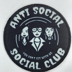 May include: A circular black patch with white text reading "ANTI SOCIAL SOCIAL CLUB" around a central graphic of three faces. The text "YOU CAN'T SIT WITH US" is below the faces, in white.