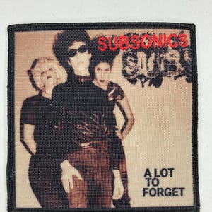 Puede incluir: Un parche de banda en blanco y negro con el texto "SUBSONICS" en rojo y "A LOT TO FORGET" en negro. El parche presenta una foto de tres personas con ropa negra.