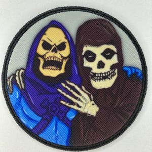 Puede incluir: Parche redondo con dos figuras de calaveras. Una calavera es amarilla con una capucha morada, la otra es marrón con una capucha negra. El parche tiene un borde negro y un fondo gris claro.