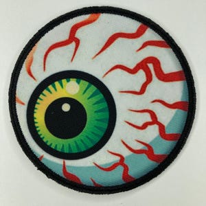Puede incluir: Un parche bordado circular con un diseño de ojo realista. El iris presenta un degradado vibrante de verde y amarillo con una pupila negra y un reflejo blanco. La esclerótica es blanca con vasos sanguíneos rojos y un borde negro.