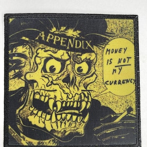 Peut inclure: Un écusson carré en tissu présentant une illustration graphique jaune et noire d'un crâne portant un chapeau. Le texte "APPENDIX" est imprimé au-dessus du crâne et une bulle contient le texte "MONEY IS NOT MY CURRENCY" en encre noire.
