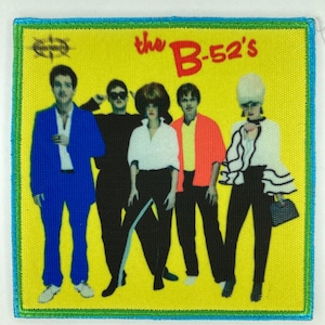 Puede incluir: Un parche bordado cuadrado con fondo amarillo y el texto "the B-52's" en rojo. El diseño muestra cinco figuras con diversos atuendos, incluyendo un traje azul, ropa negra, una camisa blanca, una chaqueta naranja y una blusa blanca con volantes.