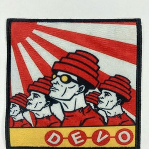 Puede incluir: Un parche bordado cuadrado con una ilustración gráfica de cuatro figuras que llevan sombreros rojos escalonados y gafas amarillas. El fondo muestra rayos de sol rojos y blancos. La palabra DEVO está impresa en letras blancas dentro de círculos rojos.