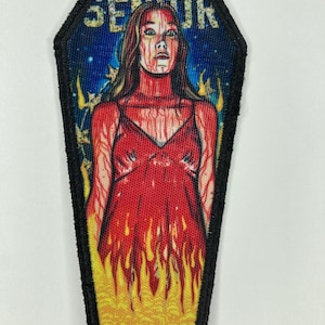 May include: A coffin-shaped embroidered patch featuring a graphic illustration of a person in a red dress covered in red liquid, standing amidst yellow and orange flames against a blue starry background, with the word SENIOR printed at the top.