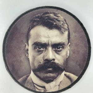 May include: Round patch featuring a sepia-toned portrait of a man with a large mustache. The image is framed by a black border. The man is wearing a collared shirt and jacket.