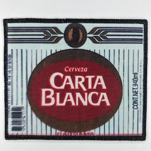 Peut inclure: Écusson rectangulaire à bordure noire. Il présente un fond bleu clair avec des lignes blanches verticales. Un motif circulaire marron et rouge au centre affiche le texte "Carta Blanca" et "Cerveza". L'écusson comprend également le texte "CONT.NET.940ml".