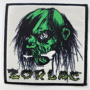 Könnte beinhalten: Quadratischer Aufnäher mit einer grünen Monstergrafik und dem Wort "ZORLAC" in grünen Buchstaben. Das Monster hat grüne Haut, schwarzes Haar und einen Knochen im Mund. Der Aufnäher hat einen schwarzen Rand.