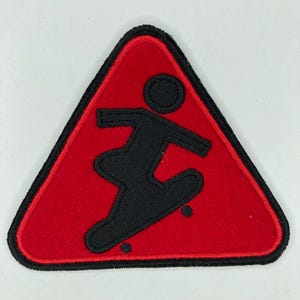 May include: Red, triangular patch with a black border and a black silhouette of a person skateboarding. The patch is a warning sign, likely for skateboarders or a skateboarding area.
