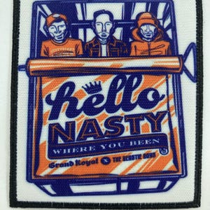 Puede incluir: Un parche bordado rectangular con borde negro que muestra tres figuras dentro de un contenedor. El texto dice hello NASTY WHERE YOU BEEN Grand Royal x THE BEASTIE BOYS en azul y naranja sobre un fondo blanco.