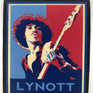 May include: A square patch featuring a stylized image of a musician playing a bass guitar. The image is divided into blue and red sections, with the name "LYNOTT" in blue text at the bottom. The patch has a black border.