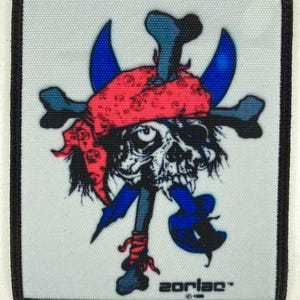 Könnte beinhalten: Quadratischer Aufnäher mit einem Totenkopf mit rotem Bandana, gekreuzten Knochen und blauen Akzenten. Der Text "zorlac" befindet sich unten. Der Aufnäher hat einen schwarzen Rand und einen weißen Hintergrund.
