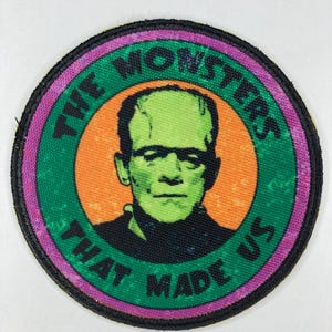 Puede incluir: Un parche redondo verde, naranja y morado con un borde negro. El parche presenta un monstruo de cara verde con el texto "THE MONSTERS THAT MADE US".