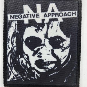 Puede incluir: Un parche bordado rectangular en blanco y negro que presenta un gráfico de alto contraste de un rostro. El texto "NA" aparece en letras blancas grandes en la parte superior, con "NEGATIVE APPROACH" impreso en una barra horizontal blanca.