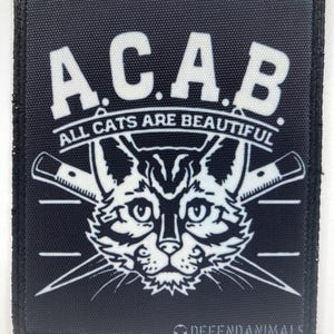 Puede incluir: Un parche rectangular negro con detalles bordados en blanco. El texto A.C.A.B. aparece en la parte superior, seguido de las palabras ALL CATS ARE BEAUTIFUL. Debajo, una cara de gato está centrada entre dos cuchillos cruzados. El texto DEFENDANIMALS está abajo.