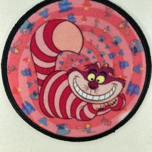 Puede incluir: Parche redondo con el Gato de Cheshire de Alicia en el país de las maravillas. El gato es de rayas rosas y rojas con una gran sonrisa y ojos amarillos. El parche tiene un borde negro y un fondo rosa con un diseño en espiral.