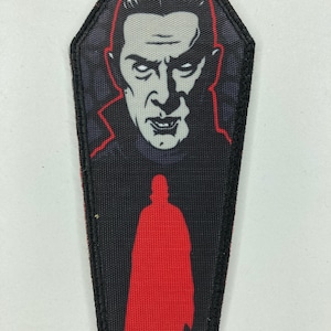 Puede incluir: Un parche negro en forma de ataúd con una ilustración roja y gris de un vampiro. La cara del vampiro está en la parte superior, y una silueta roja de una figura con capa está debajo. El parche tiene un borde negro.