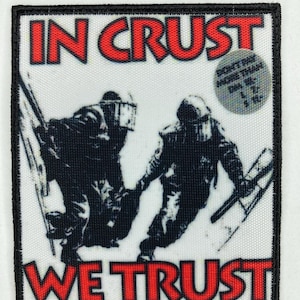 Peut inclure: Un patch noir et blanc avec les mots "In Crust We Trust" en lettres rouges. Le patch présente une image en noir et blanc de deux personnages portant des équipements anti-émeute.