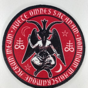 Puede incluir: Parche redondo rojo con borde negro. Presenta una ilustración negra de Baphomet, una deidad con cabeza de cabra, con alas y postura sentada. Texto blanco y símbolos, incluidas estrellas y lunas crecientes, en el borde.