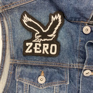 Könnte beinhalten: Eine blaue Jeansjacke mit einem schwarz-weißen Aufnäher, der einen stilisierten Adler mit ausgebreiteten Flügeln und dem Wort "ZERO" zeigt. Die Jacke hat silberne Knöpfe und zwei Vordertaschen.