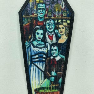 Könnte beinhalten: Ein Sarg-förmiger Aufnäher mit einem Familienporträt der klassischen Monsterfamilie. Das farbenfrohe Bild zeigt Frankensteins Monster, einen Vampir und andere Charaktere. Der Aufnäher hat einen schwarzen Rand.