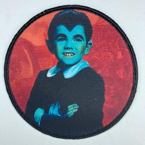 Könnte beinhalten: Ein runder, schwarz umrandeter Patch mit einem bunten Bild eines jungen Jungen mit einem schwarzen Kurzhaarschnitt, der ein schwarzes Hemd mit Kragen und einen weißen Kragen trägt. Der Junge hat ein blaues Gesicht und lächelt. Der Hintergrund ist rot und orange.