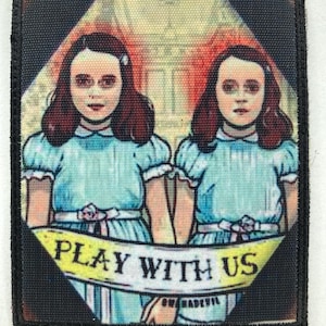 Könnte beinhalten: Quadratischer Aufnäher mit schwarzem Rand, der zwei Mädchen in blauen Kleidern zeigt, die sich an den Händen halten. Ein gelbes Banner am unteren Rand trägt die Aufschrift "PLAY WITH US". Der Hintergrund zeigt ein Gebäude in Rot- und Gelbtönen.