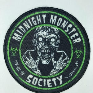 Könnte beinhalten: Ein schwarzer und grüner runder Patch mit einem weißen Zombiegesicht in der Mitte. Der Text "Midnight Monster Society" ist in einem weißen Kreis um das Zombiegesicht geschrieben.