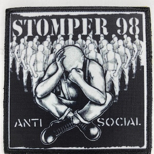 Op de afbeelding: Zwart-wit patch met de tekst "STOMPER 98" boven een illustratie van een persoon met zijn hoofd in zijn handen, omringd door een menigte. De woorden "ANTI SOCIAL" staan eronder.