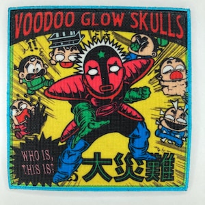 May include: A square embroidered patch featuring a central masked figure in red and black with green accents, surrounded by smaller cartoon characters on a yellow background. The text reads VOODOO GLOW SKULLS, WHO IS, THIS IS?, and Japanese characters.