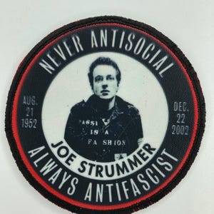 May include: A circular embroidered patch featuring a black and white portrait of Joe Strummer. The text reads NEVER ANTISOCIAL, ALWAYS ANTIFASCIST, JOE STRUMMER, AUG 21 1952, and DEC 22 2002. The patch has a black border with a thin red inner ring.