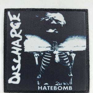 Pode incluir: Etiqueta bordada preta e branca com o texto "DISCHARGE" e "HATEBOMB" em um fundo preto. A etiqueta apresenta uma imagem de esqueleto.