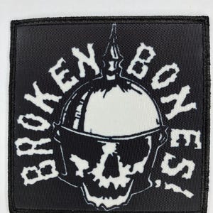 May include: Black square patch with white text and a skull design. The text reads "BROKEN BONES!" in a stylized font, surrounding a skull wearing a helmet. The patch has a black border.