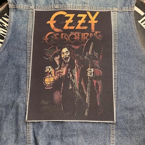 Puede incluir: Un chaleco de mezclilla azul con un parche de tela rectangular grande cosido en la parte posterior. El parche muestra el texto Ozzy Osbourne en letras naranjas sobre un gráfico de una figura que sostiene una linterna encendida y una pala.