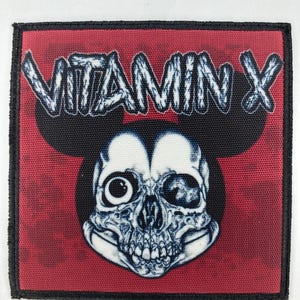 May include: A red fabric patch with black stitching and a white skull with one eye. The skull is wearing Mickey Mouse ears. The text "VITAMIN X" is written in white on a black background.