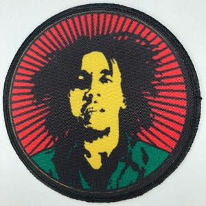 May include: A round black patch with a red, yellow, and green image of Bob Marley. The image is surrounded by a red sunburst.