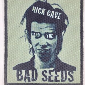 May include: A rectangular fabric patch featuring a screen-printed portrait of Nick Cave. The design uses a muted sage green and dark charcoal color palette. The text NICK CAVE AND THE BAD SEEDS is printed in a bold, sans-serif font across the image.
