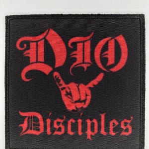 Puede incluir: Parche cuadrado negro con texto rojo y un gesto con la mano. El texto dice "DIO" encima de "Disciples" en una fuente gótica. El gesto de la mano es un dedo índice y meñique levantados.